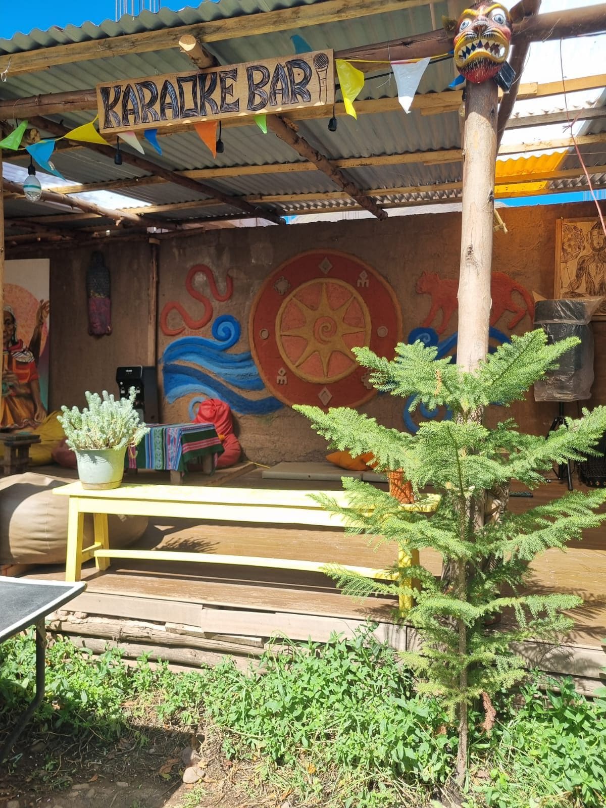 Un área de estar al aire libre rústica con una mesa y sillas de madera, una planta pequeña y un cartel que dice 'KARALIKE BAR'.
