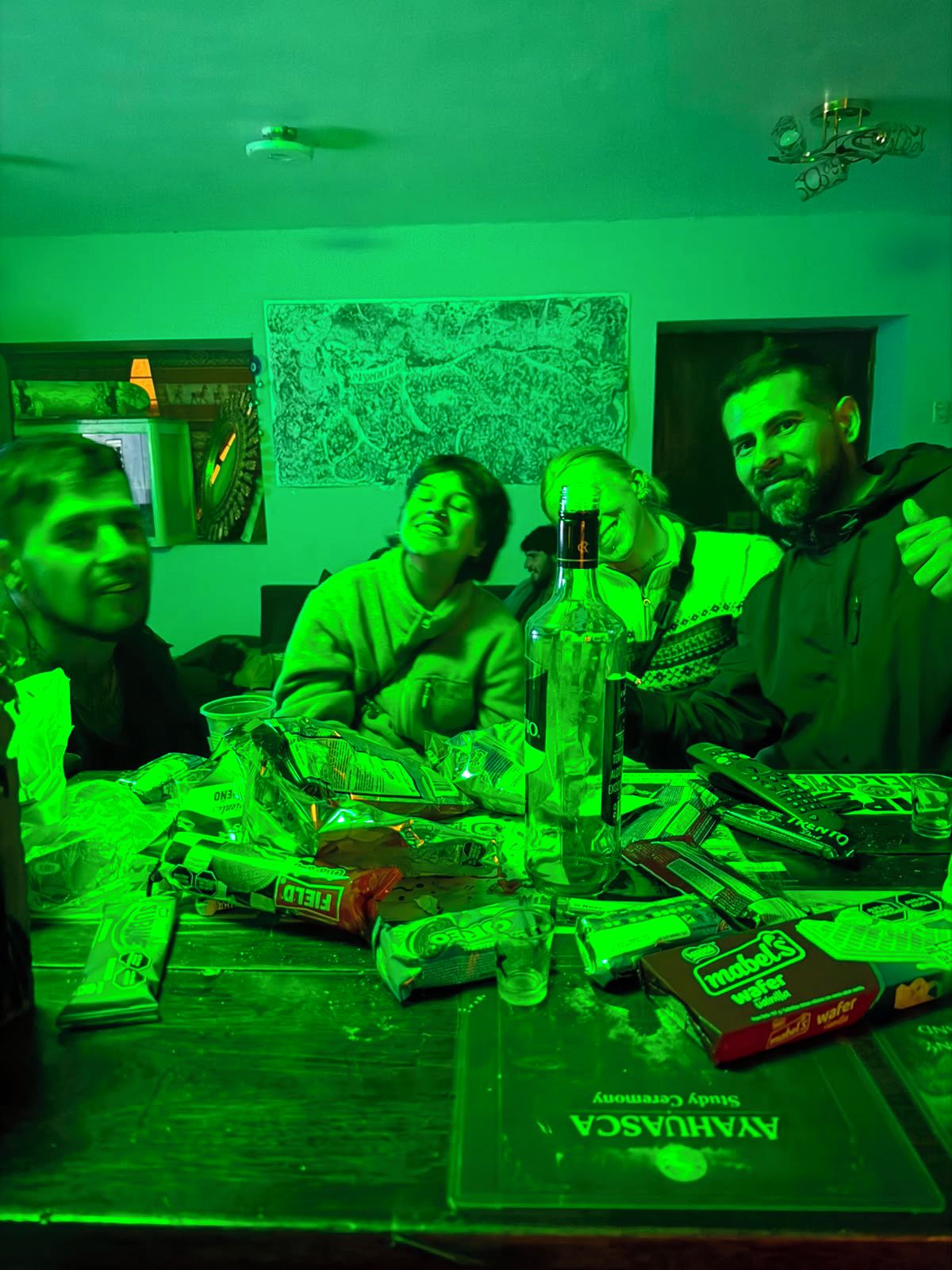Un grupo de personas alrededor de una mesa con diversos objetos, incluyendo una botella y cajas, en una habitación con un tono verdoso.