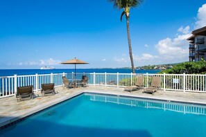 A pool area with a view of the ocean, a palm tree, and a white fence. - Kona Shores#223: Top Floor, Split AC, Remodeled! (Kailua-Kona)