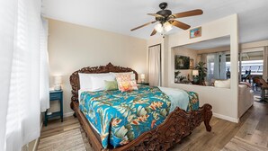 Un dormitorio con una cama grande, un ventilador de techo, una mesita de noche, una ventana con vistas y una puerta que da a otra habitación.