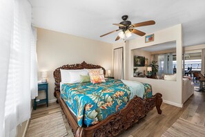 A bedroom with a large bed, a ceiling fan, a nightstand, a window with a view, and a door leading to another room. - Kona Shores#223: Top Floor, Split AC, Remodeled! (Kailua-Kona)