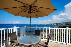 A balcony with a table and chairs, a large umbrella, and a view of the ocean and a cruise ship. - Kona Shores#223: Top Floor, Split AC, Remodeled! (Kailua-Kona)