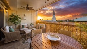 Un balcón con mobiliario de mimbre, una mesa y vistas a la aguja de una iglesia al atardecer.