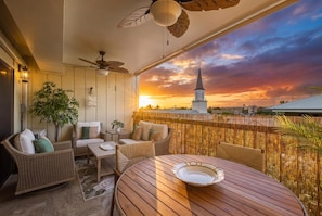 A balcony with wicker furniture, a table, and a view of a church spire during sunset. - Kona Plaza#417: Top Floor, Downtown! (Kailua-Kona)