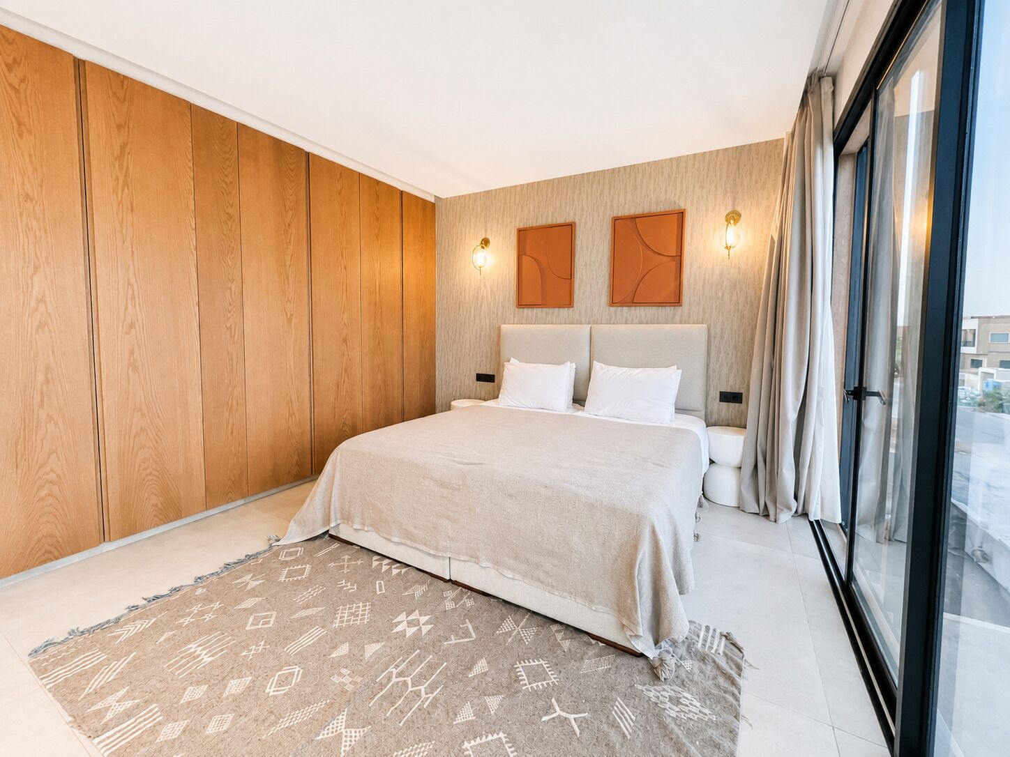 A modern bedroom with a large bed, wooden panel walls, a patterned rug, and a view through a large window.
