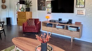 Een woonkamer met een rode fauteuil, een houten salontafel, een aan de muur bevestigde televisie en een boekenkast met boeken en decoratieve objecten.