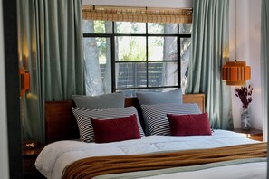 Un dormitorio con cama, dos almohadas, una mesita de noche con lámpara y una ventana con cortinas.