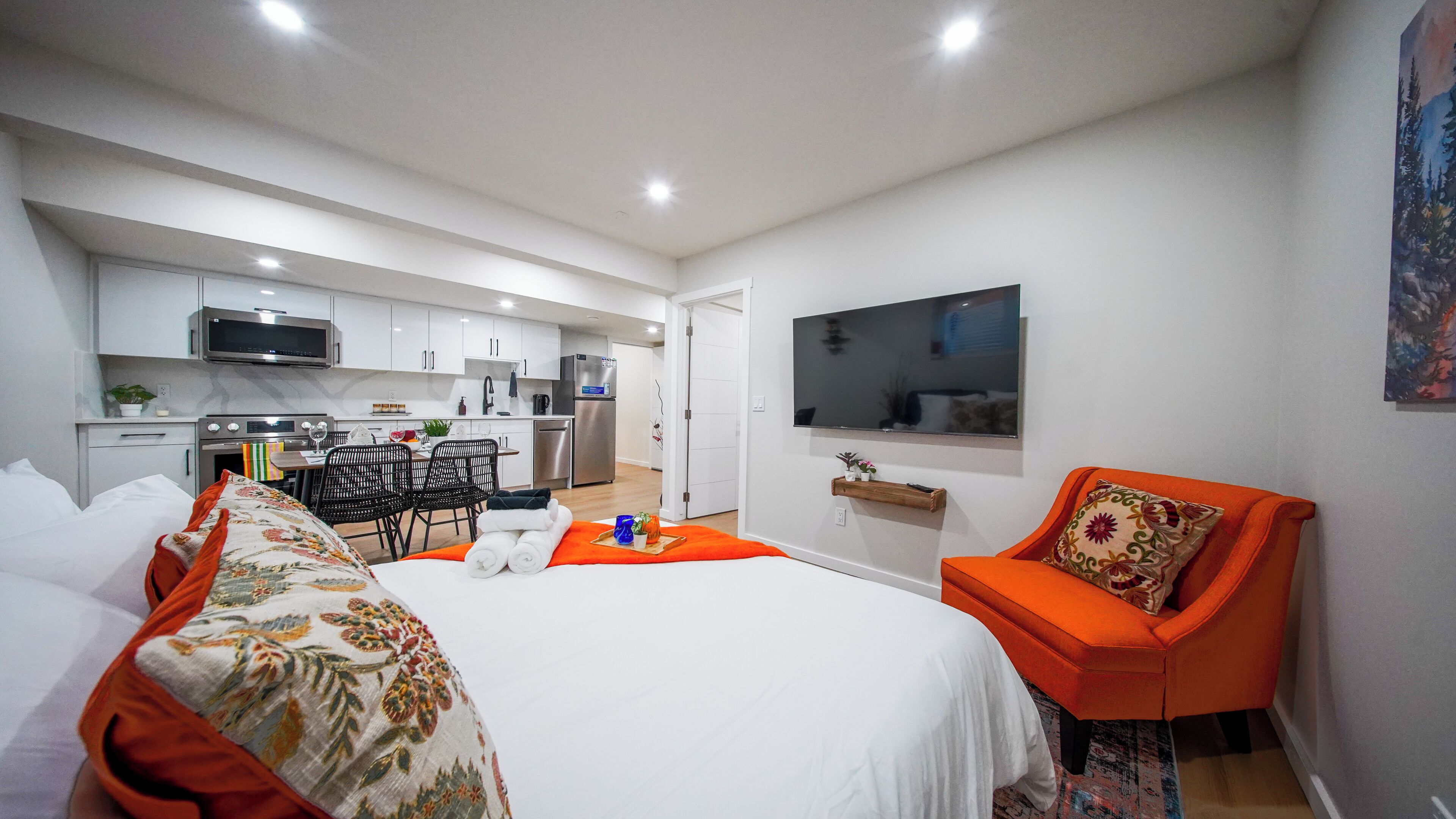 Habitación de hotel con cama, zona de estar con una silla, una pequeña mesa, televisor montado en la pared y una kitchenette al fondo.