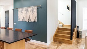 A modern kitchen with a dark teal accent wall, a wooden dining table with brown chairs, and a staircase with wooden steps and a black railing.