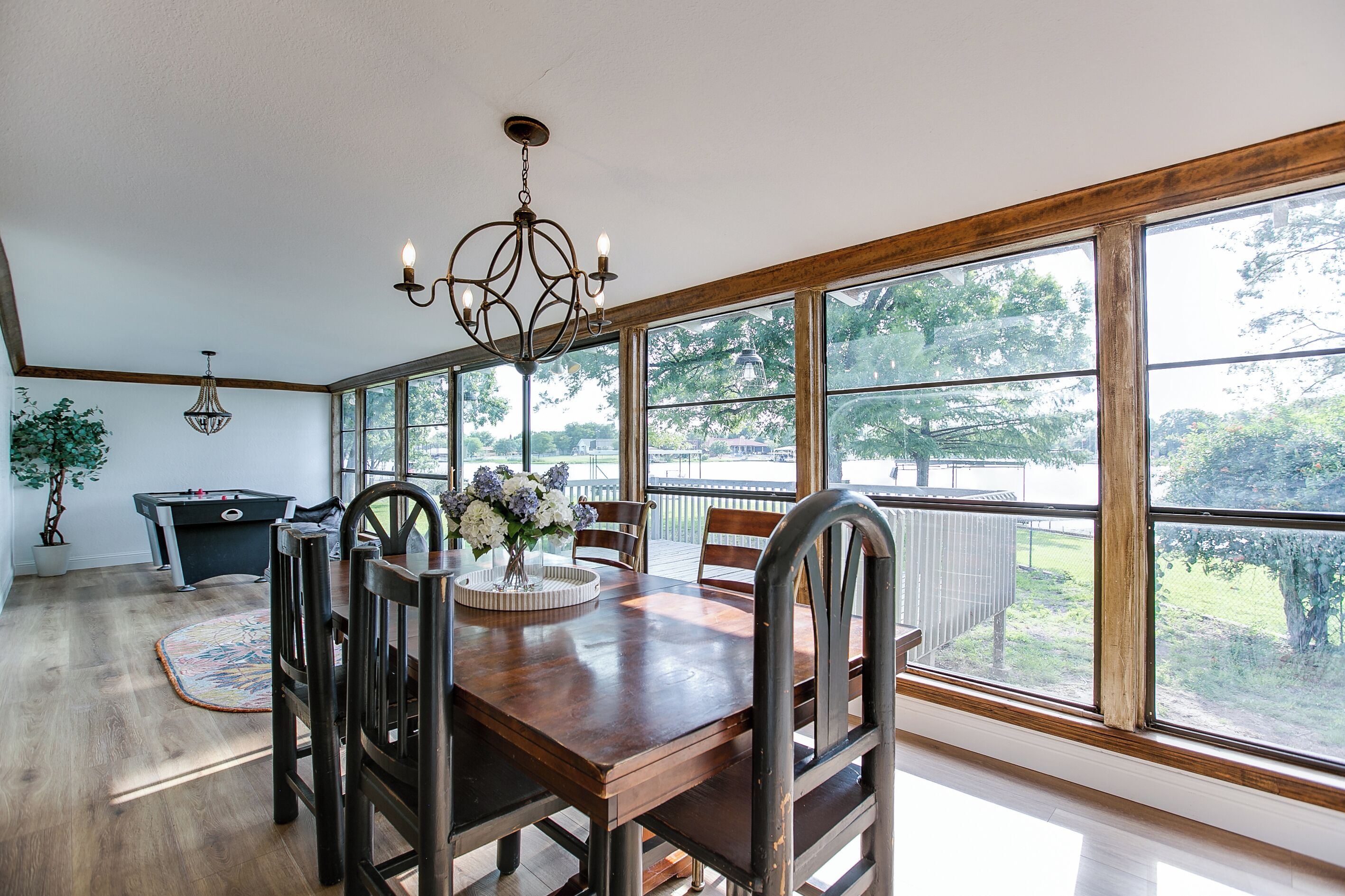 A dining room with a wooden table, chairs, a chandelier, and large windows offering a view of the outdoors.