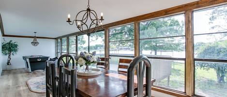 A dining room with a wooden table, chairs, a chandelier, and large windows offering a view of the outdoors.