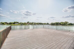 A wooden deck with a metal railing overlooking a body of water and a park with trees and buildings in the background. - Lakeside Home w/Private Casita & Stunning Views (San Angelo)