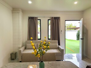 A modern living room with a beige sofa, a vase with yellow flowers, and a view of a green lawn through a door. - 3-bedroom house in adorable Chalong Phuket with nestled among lush mature trees (Tambon Chalong)