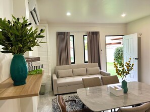 Una sala moderna con un sofá, una mesa de comedor y una mesa de centro. Hay una planta a la izquierda y una ventana con cortinas.