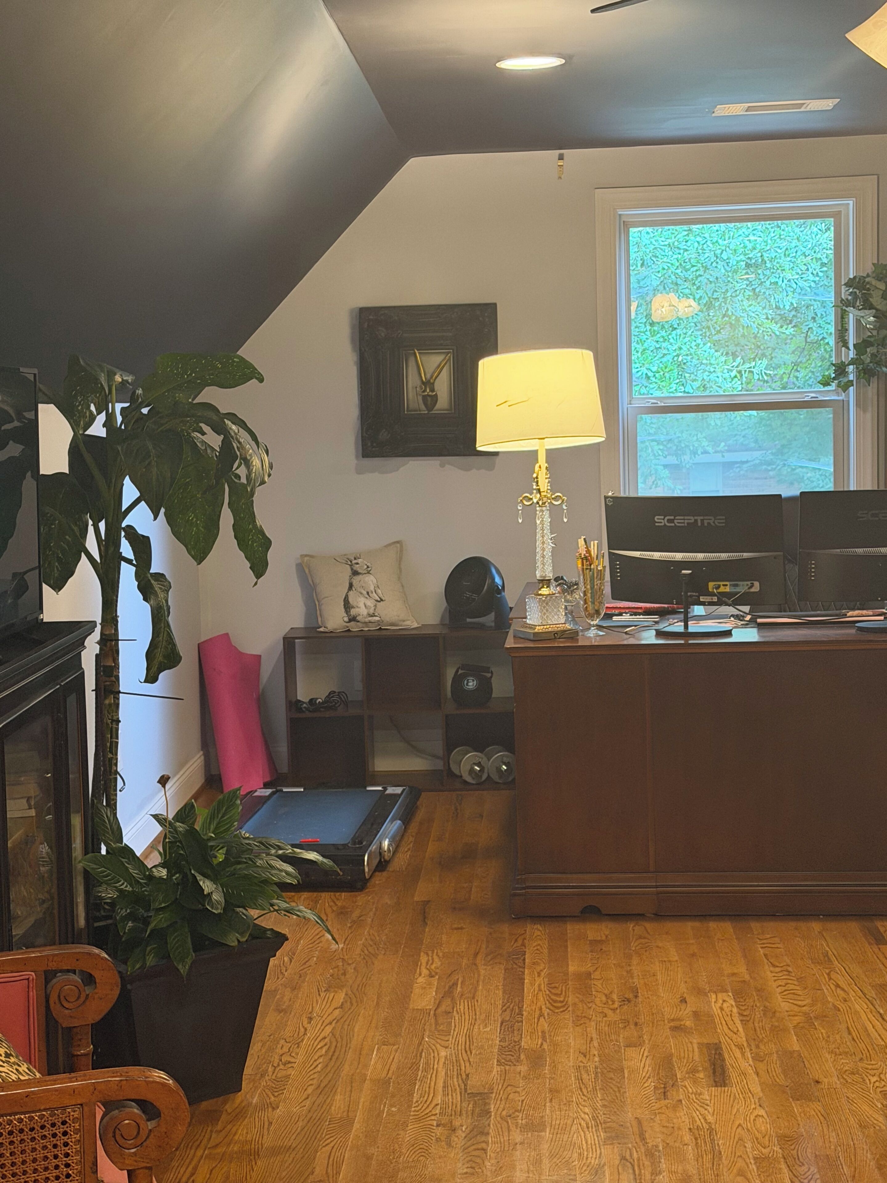 A room with a wooden floor, a desk with a computer, a lamp, and a large window with a view of greenery.