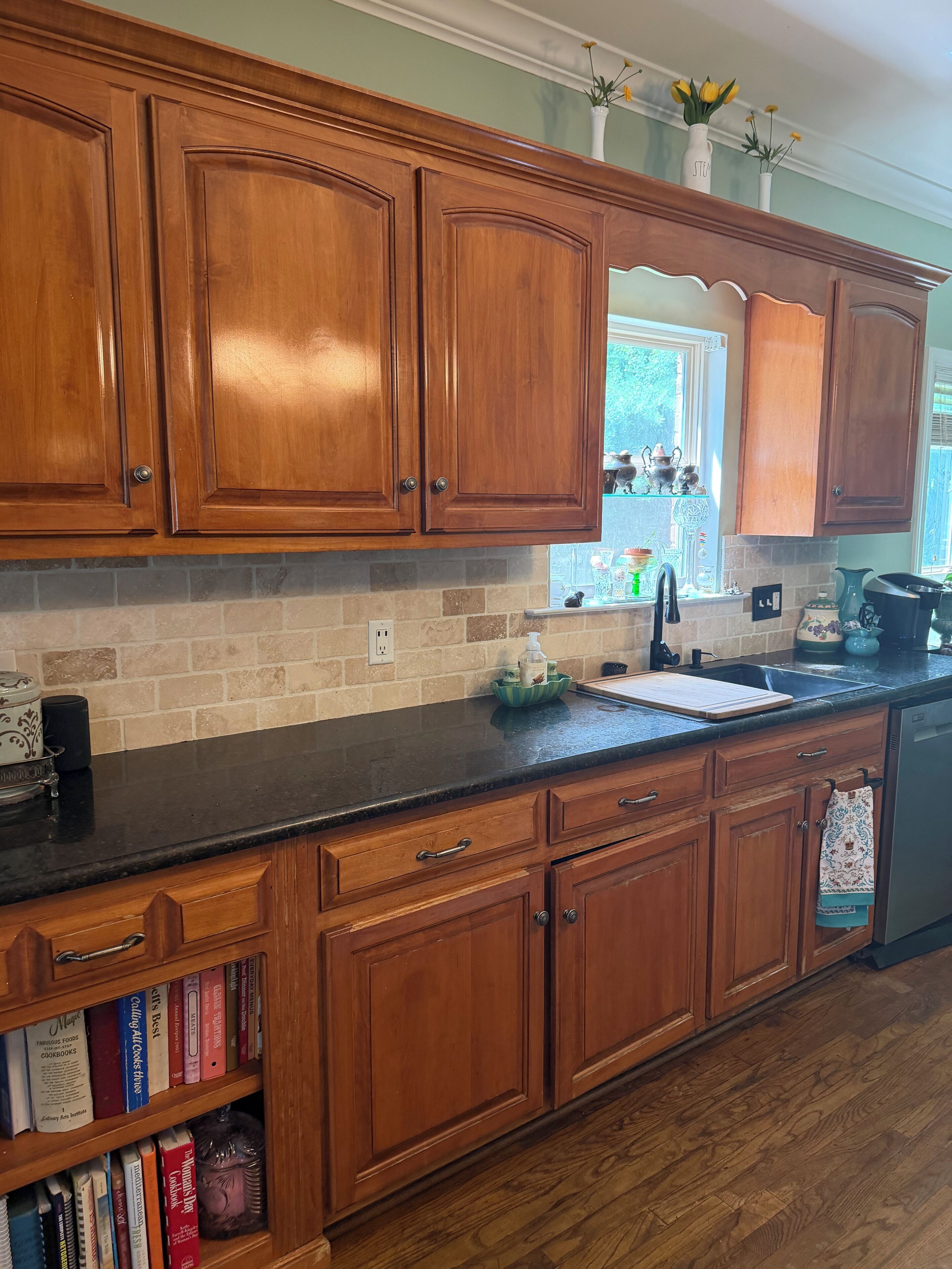 A kitchen with wooden cabinets, a black countertop, a built-in bookshelf, and a window with decorative items.