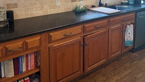 A kitchen with wooden cabinets, a black countertop, a built-in bookshelf, and a window with decorative items.