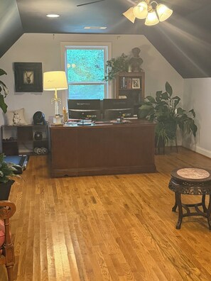 A room with a wooden floor, a desk with two monitors, a lamp, and a ceiling fan.
