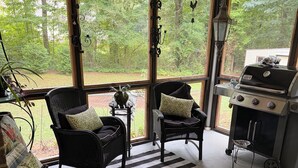 A covered porch with a grill, two chairs, a striped rug, and a view of the yard.