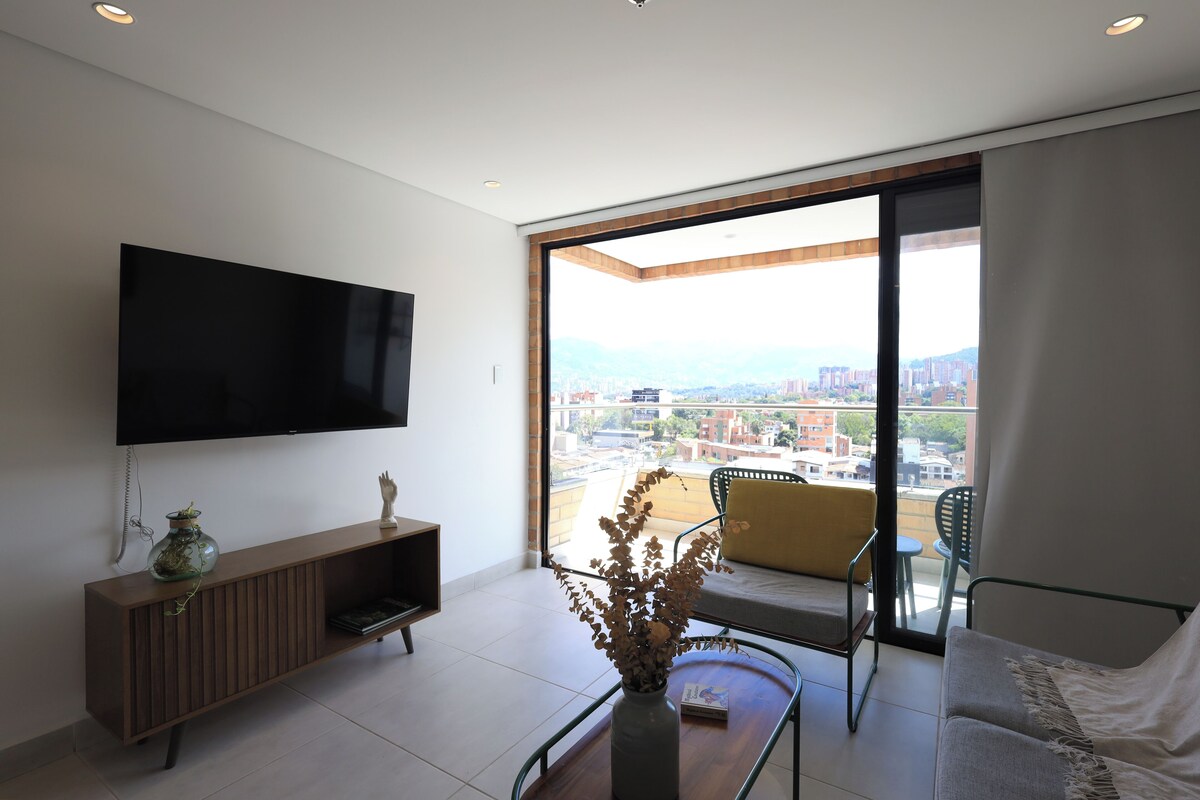 A modern living room with a flat-screen TV, a wooden console, a plant, a sofa, and a view of a cityscape through large glass doors.