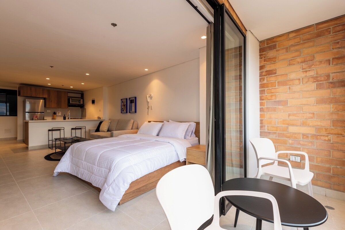 A modern hotel room with a bed, a dining table, and a kitchen area.