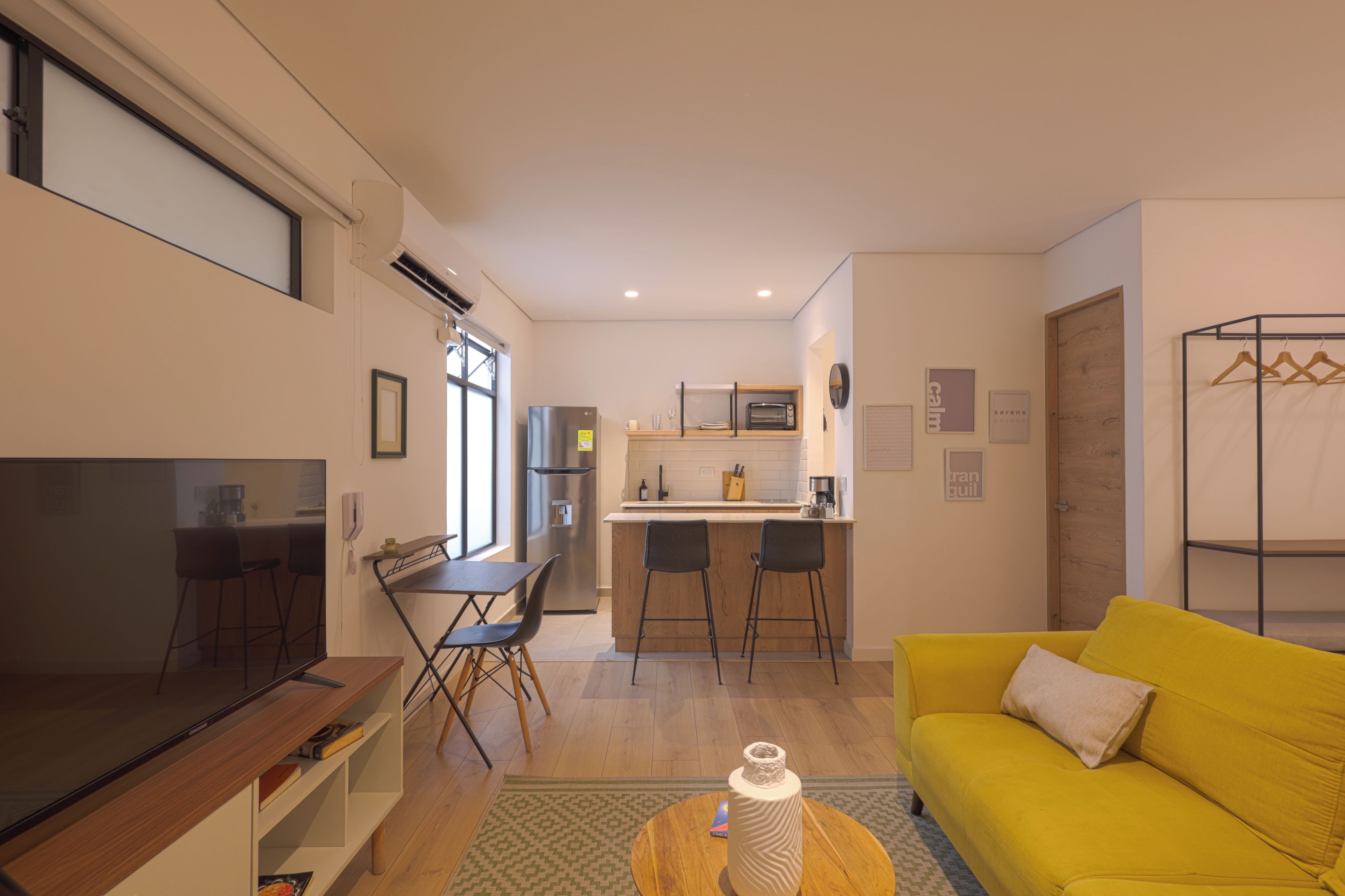 A modern living room with a flat-screen TV, a yellow sofa, and a kitchen area with a refrigerator and microwave.