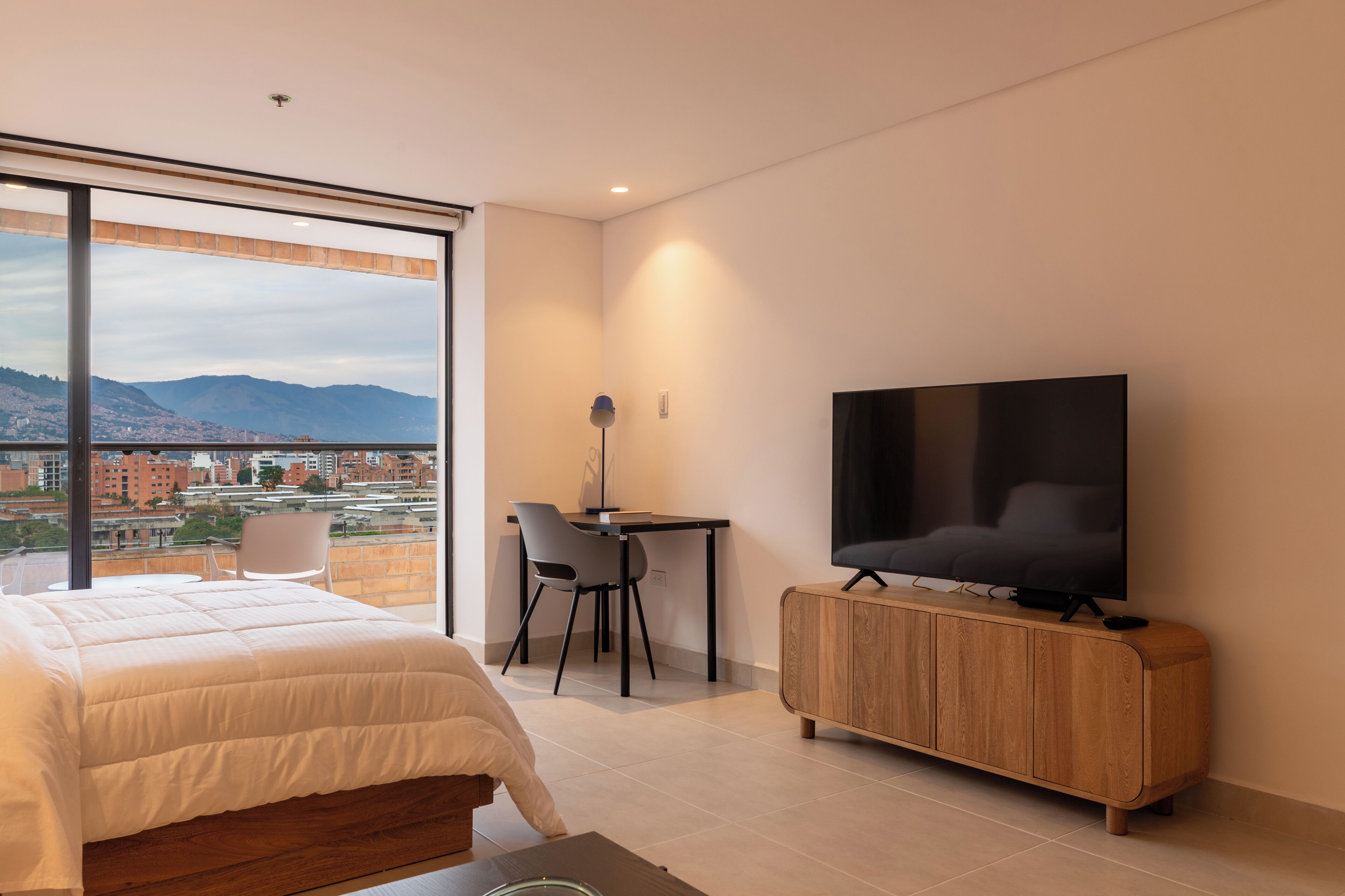 A modern hotel room with a large bed, a desk with a chair, a television, and a view of a cityscape through large windows.