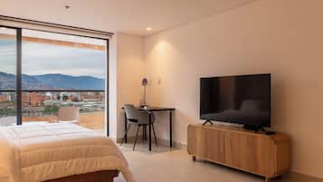 A modern hotel room with a large bed, a desk with a chair, a television, and a view of a cityscape through large windows.