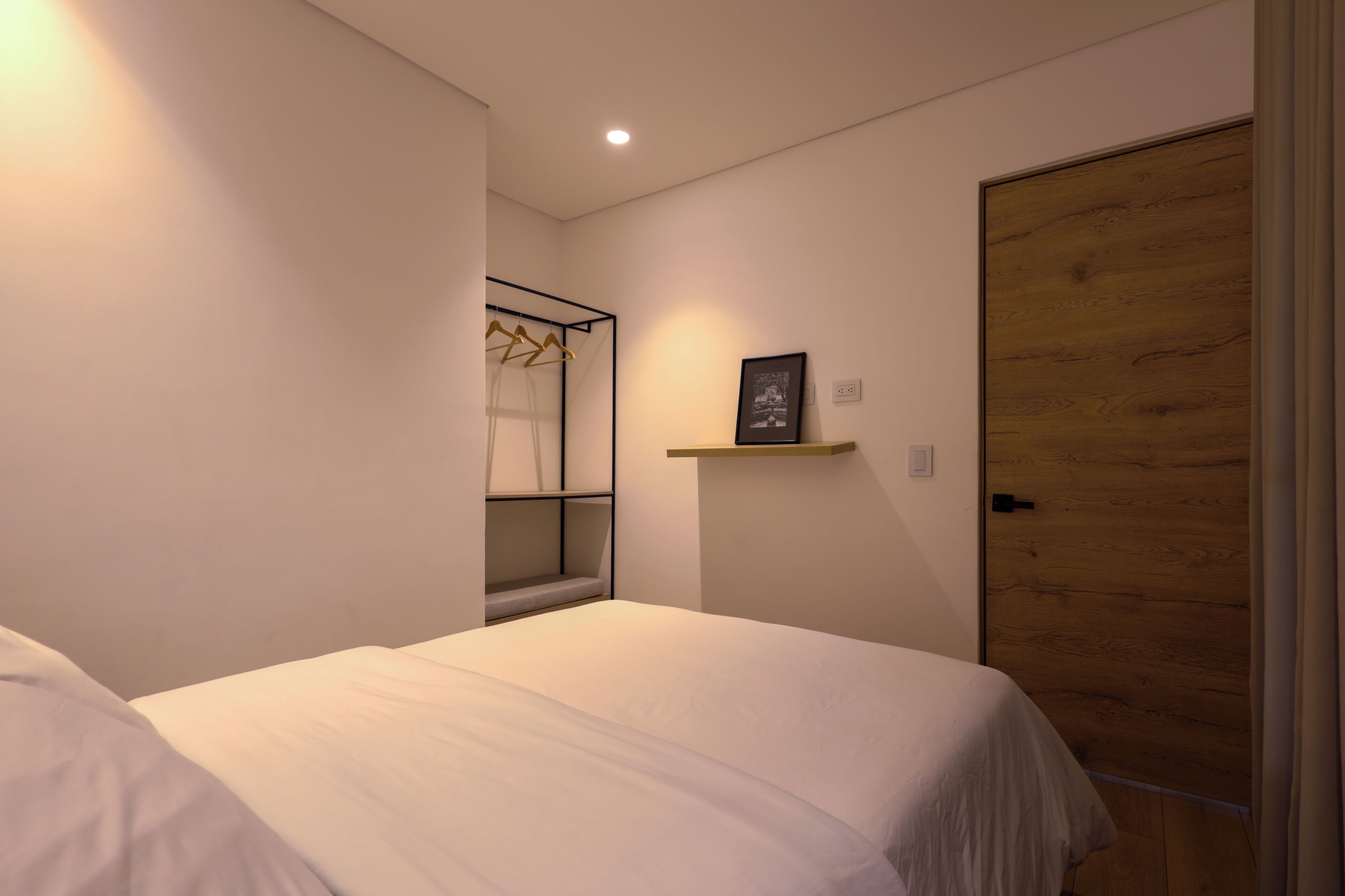 A bedroom with a bed, a wooden door, a wall-mounted shelf with a picture, and a clothing rack.