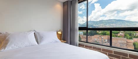 A hotel room with a large bed, a nightstand, a lamp, and a window with a city view.