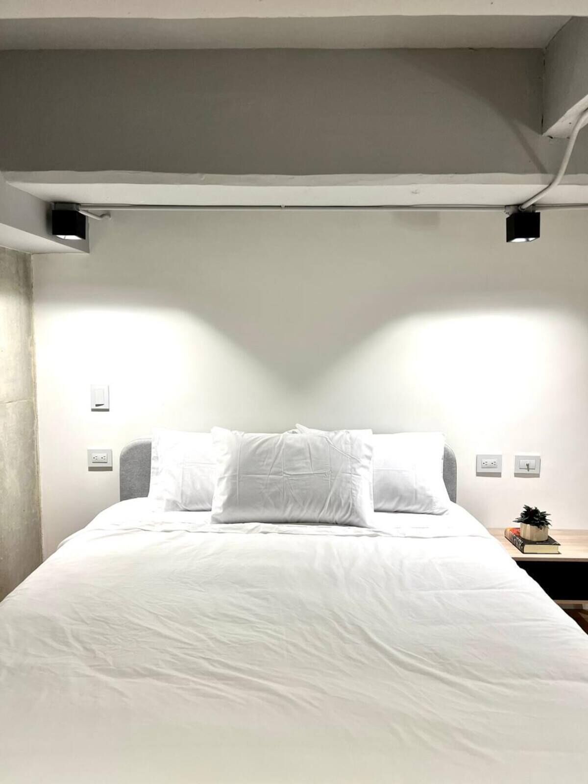 Ein Bett mit weißer Bettwäsche, ein Nachttisch mit einem Buch und einer kleinen Pflanze sowie eine Leuchte über dem Bett.