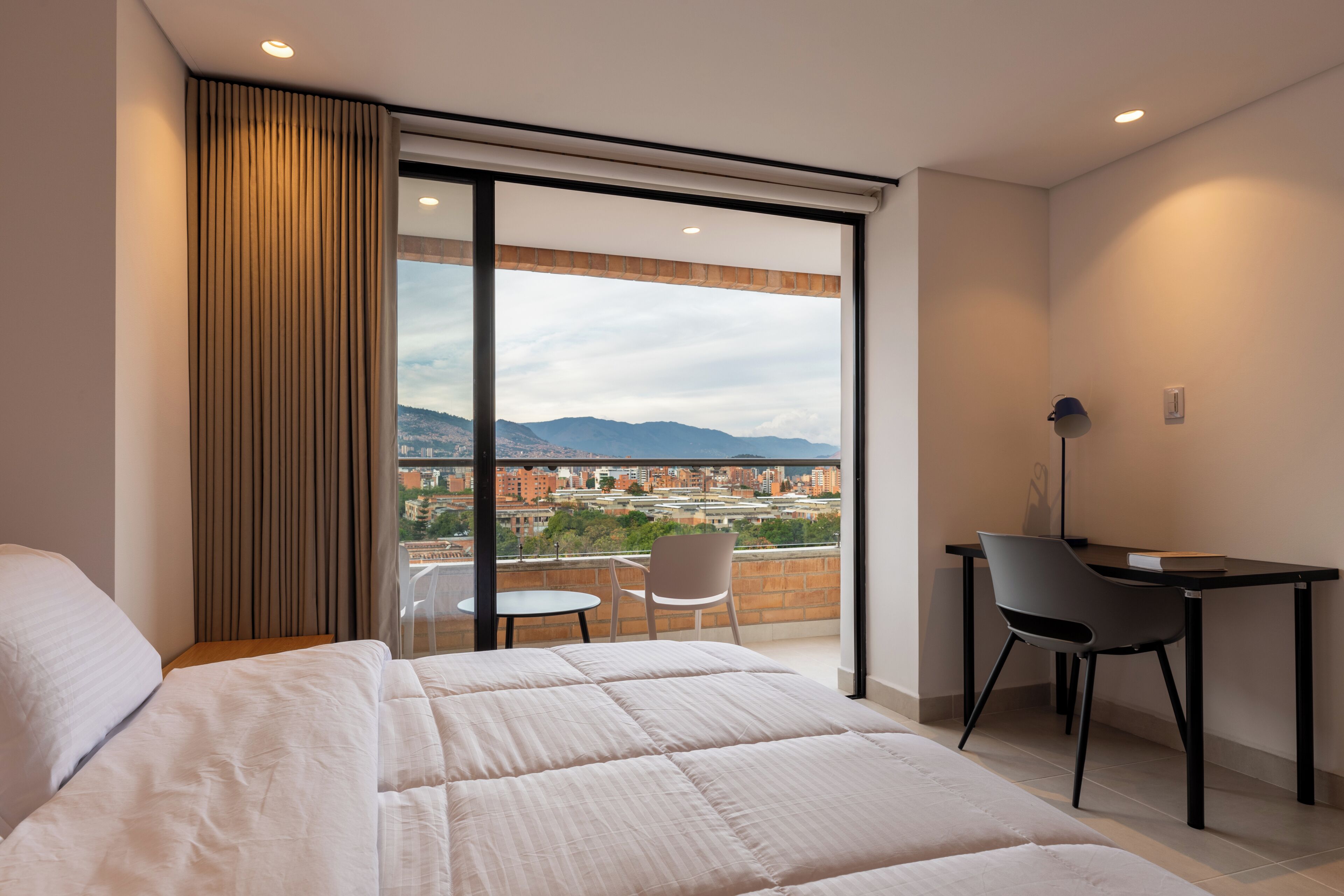 A hotel room with a bed, a desk, and a chair. There is a large window with a view of a cityscape and mountains.