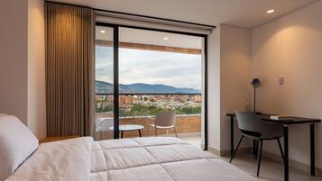 A hotel room with a bed, a desk, and a chair. There is a large window with a view of a cityscape and mountains.