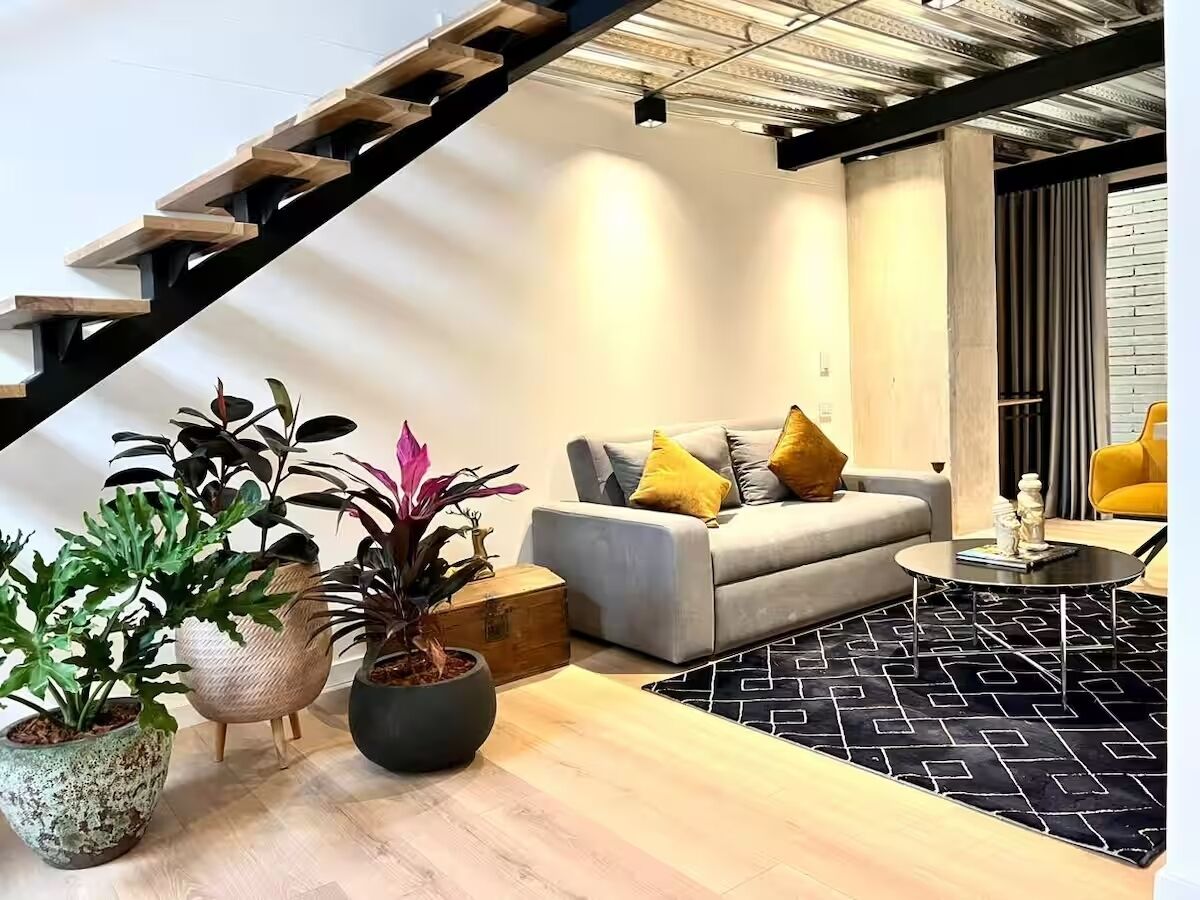 Un espacio moderno con escalera, un sofá gris con cojines amarillos y plantas en macetas.