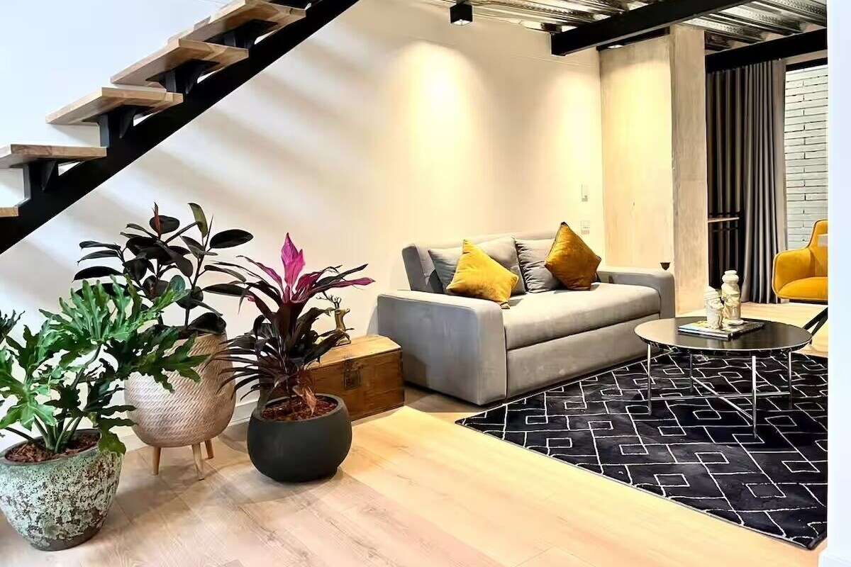A modern living space with a staircase, a grey sofa with yellow cushions, and potted plants.