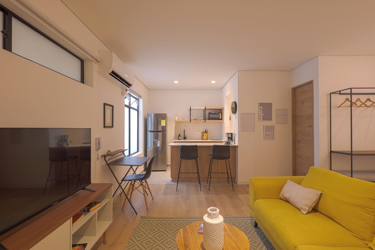 A modern living room with a flat-screen TV, a yellow sofa, and a kitchen area with a refrigerator and microwave.