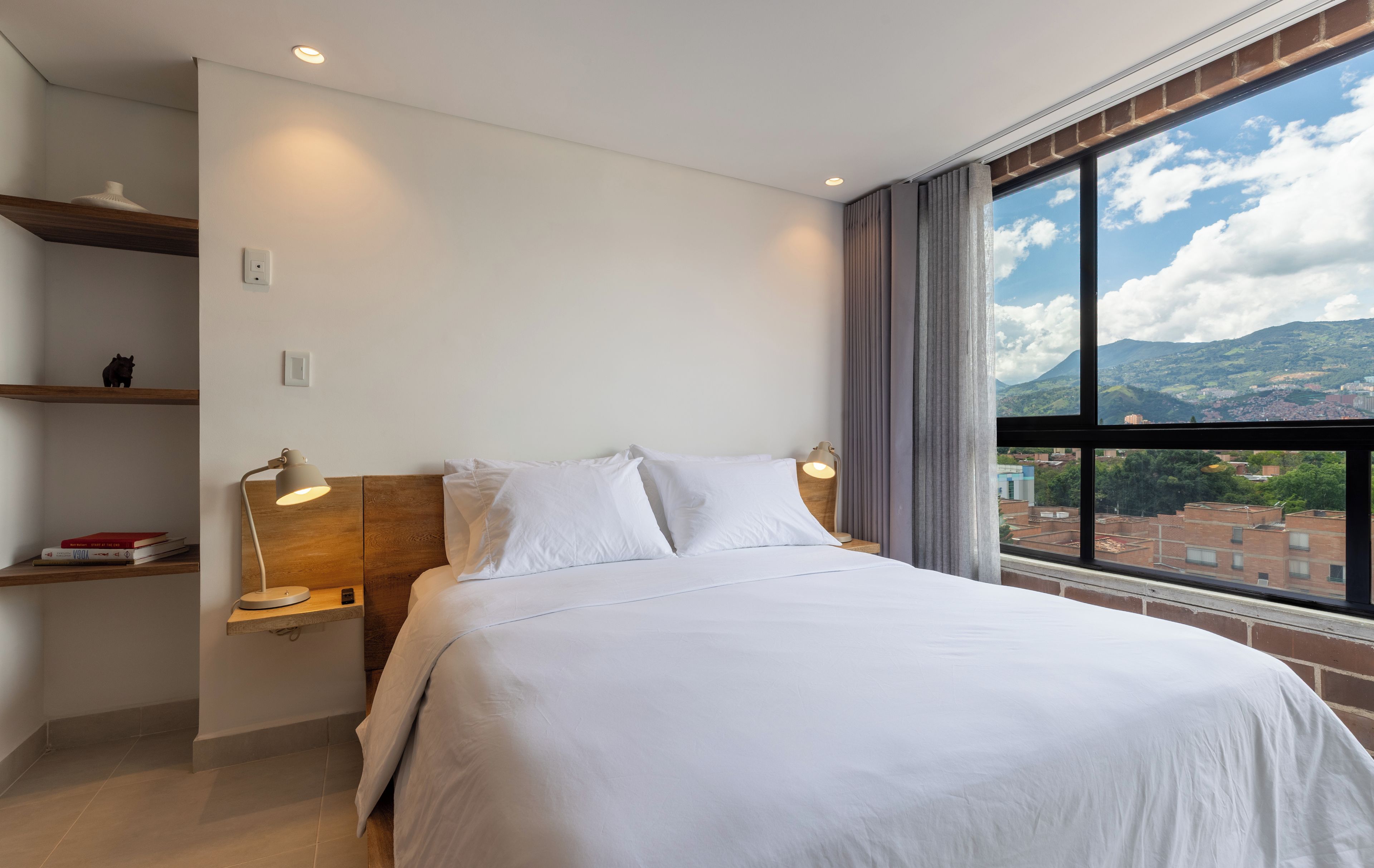 Una habitación de hotel moderna con una cama grande, una mesita de noche, una estantería con libros y un ventanal con vistas a la ciudad.