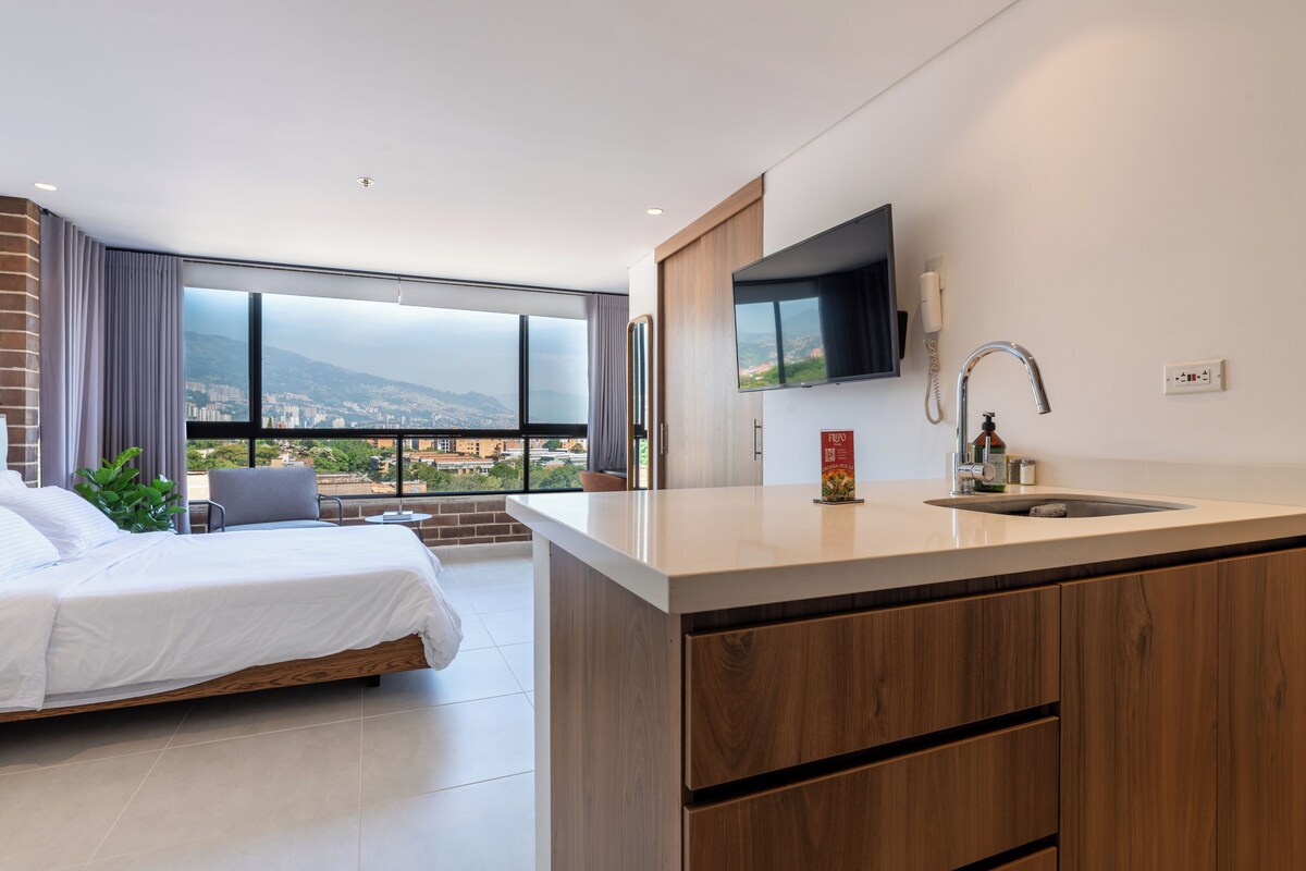 A modern hotel room with a large bed, a small seating area, a kitchen with a sink, and a TV mounted on the wall.