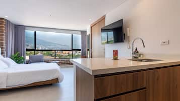 A modern hotel room with a large bed, a small seating area, a kitchen with a sink, and a TV mounted on the wall.