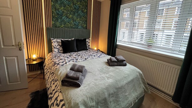 A hotel room with a bed, a nightstand, a wall-mounted headboard, a window with blinds, and a door. - Spacious & Modern Family Gem (London)