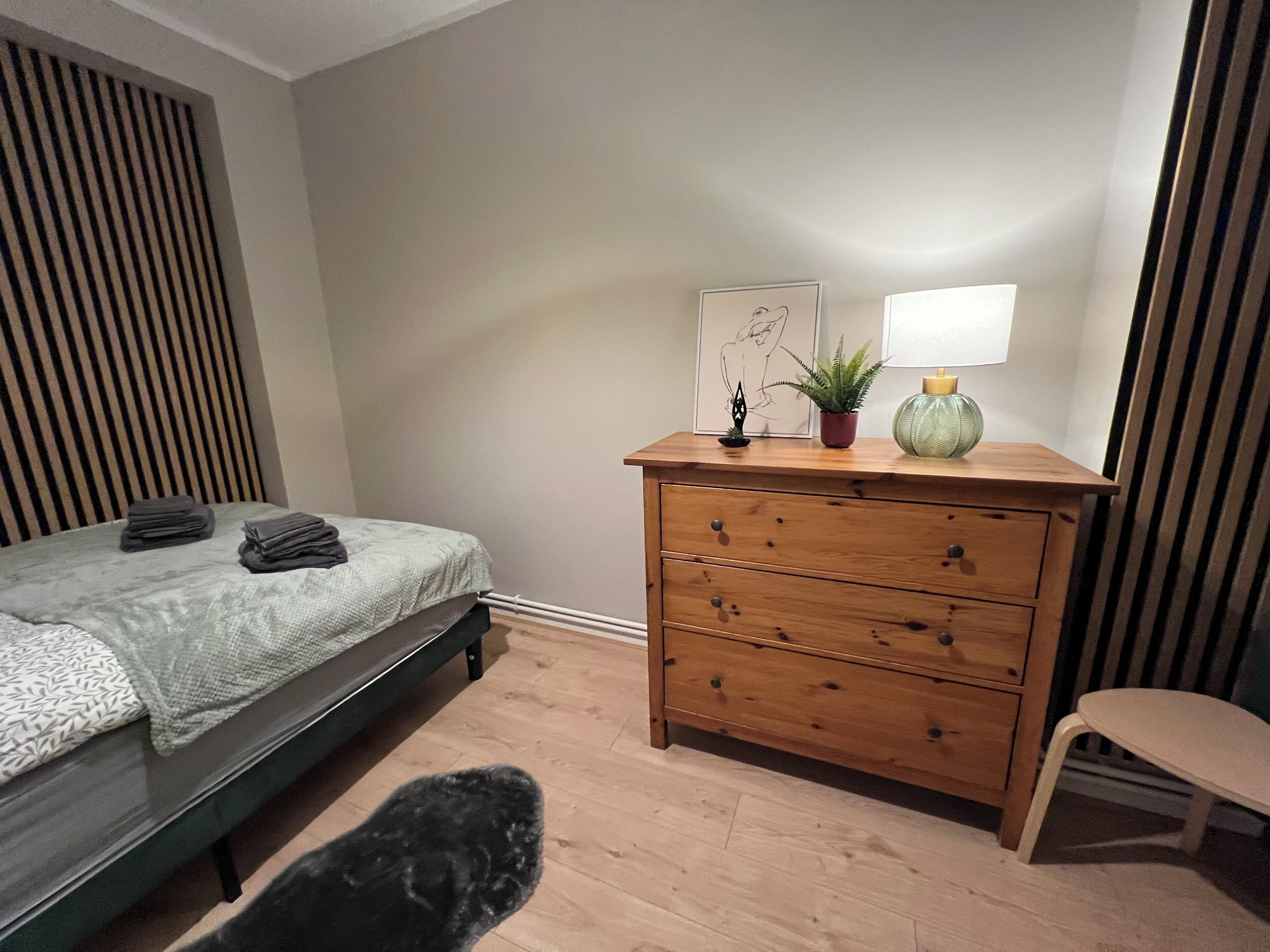 Une chambre à coucher avec un lit, une commode en bois, une chaise et une table de chevet sur laquelle se trouvent une lampe et une plante.