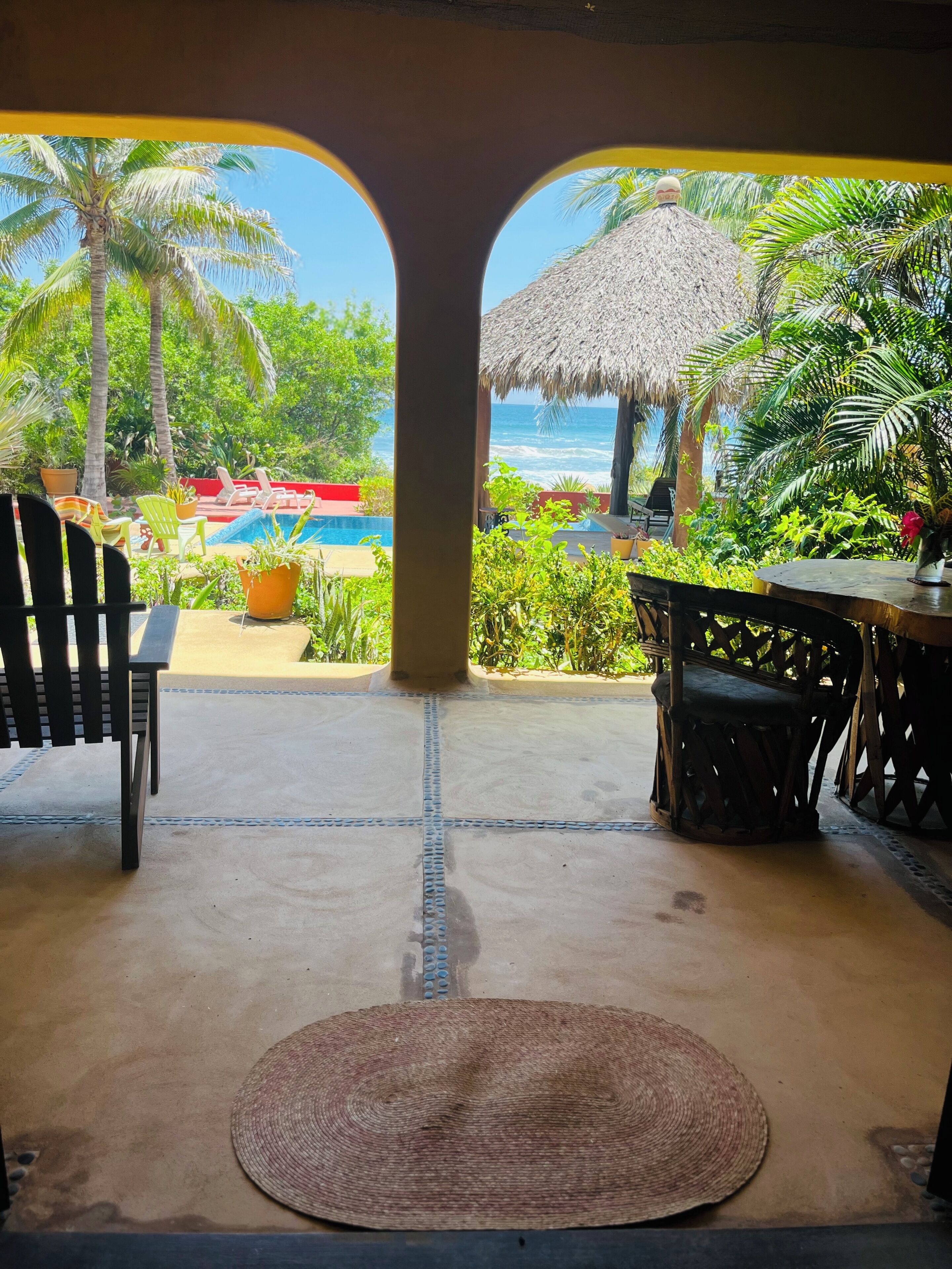 A view from a covered area with a thatched roof, overlooking a pool and a beach with palm trees.