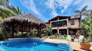 Zona de piscina con un pabellón de techo de paja, un edificio de dos pisos con balcón y vegetación tropical.