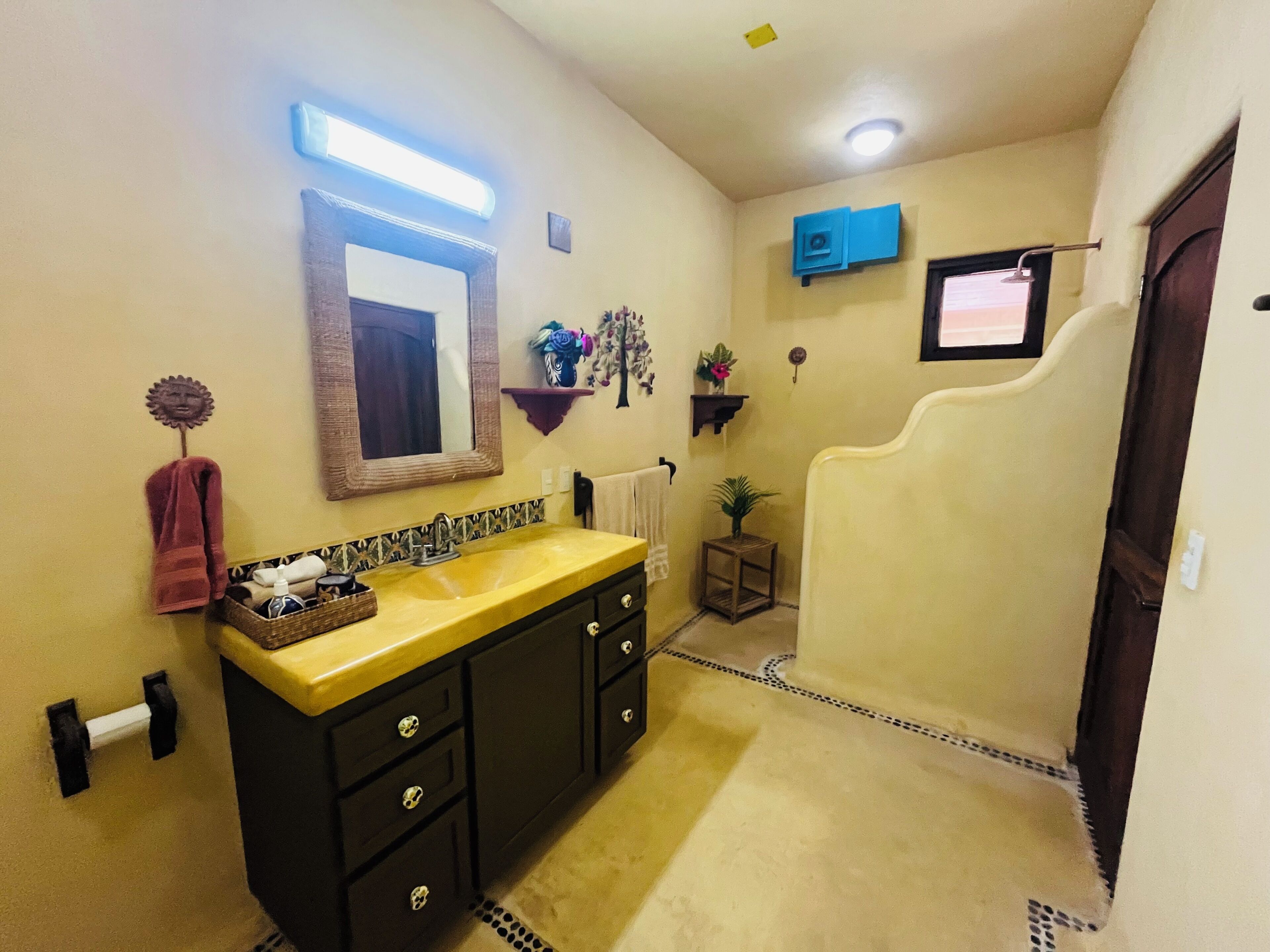 A bathroom with a yellow sink, dark cabinets, a mirror, a towel rack, a small shelf with flowers, and a shower area.