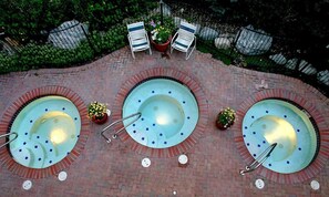 Ein Poolbereich auf dem Dach mit drei Whirlpools, umgeben von Pflastersteinen und zwei Liegestühlen mit Topfpflanzen.