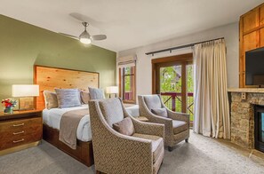 Ein Schlafzimmer mit einem Bett, zwei Sesseln, einem Holz-Kopfteil, einem Nachttisch, einer Lampe, einem Fernseher, einem Steinkamin und einem Fenster mit Vorhängen.