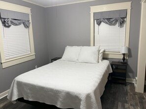 Un dormitorio con cama, dos mesitas de noche, lámpara y ventanas con persianas.
