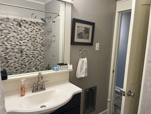 Un baño con un lavabo blanco, una ducha con puerta de cristal, un cuadro enmarcado en la pared y una toalla colgada en un perchero.
