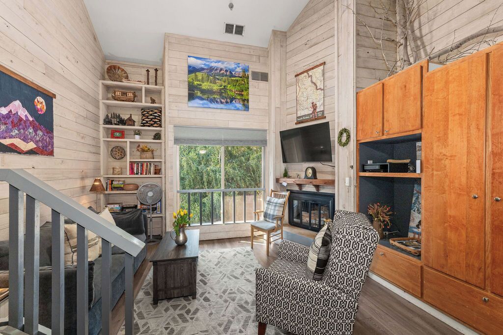 A spacious living room with a staircase, wooden walls, a large window, and a fireplace.