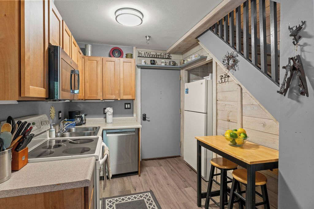 A kitchen with wooden cabinets, a stainless steel refrigerator, a dishwasher, and a wooden dining table with two stools.